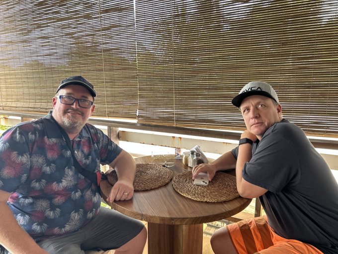 Two men sitting at a round wooden table in what appears to be a bamboo-walled restaurant or café; the man on the left wears glasses and a black cap with a patterned shirt, while the man on the right wears a gray cap and dark shirt with orange shorts.