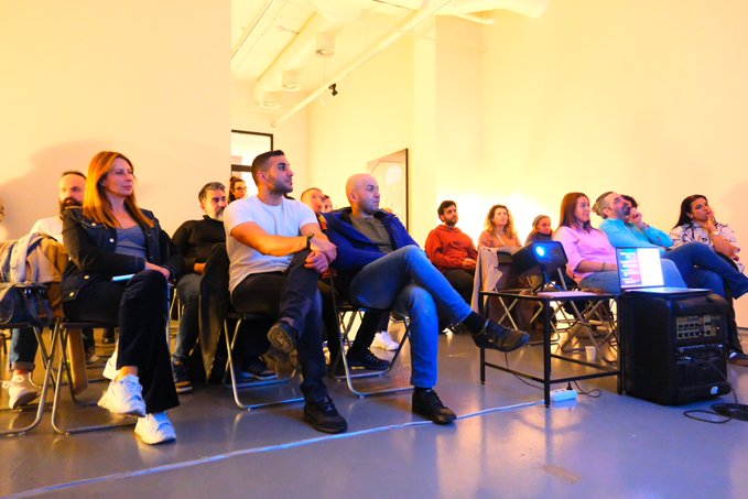 An audience seated indoors watches attentively, with a projector visible in the foreground and attendees including a man in a blue blazer and a woman in a black blazer.