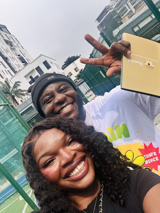 A cheerful selfie captures a smiling man and woman outdoors near a tennis court, with the man holding a gold box labeled 'Haeden' and making a peace sign.