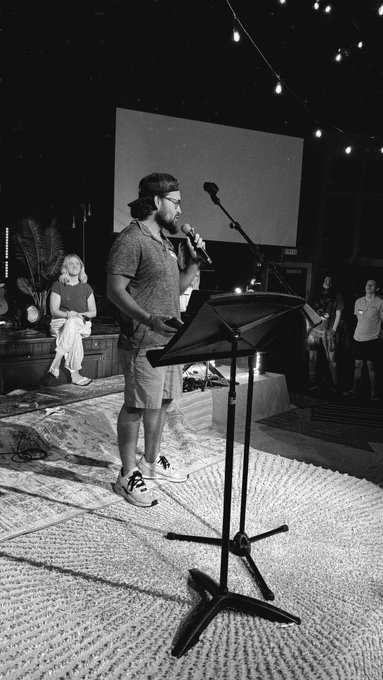 A bearded man in a cap and shorts speaks into a microphone at a music stand on a rug-covered stage, with a projector screen and string lights behind him, capturing an intimate community gathering that fits the rallying "Good Morning Degens & Gamers - TODAY IS OUR OPPORTUNITY" message.
