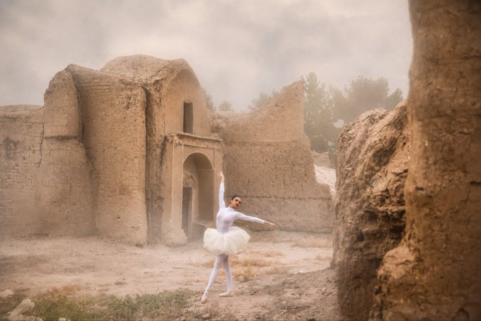 A dancer in a white tutu and tights performs a graceful pose amidst ancient, weathered earthen ruins under a cloudy sky.