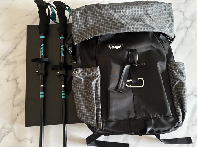 A gray and black Bitget-branded backpack lies on a marble surface next to two black and teal trekking poles, also branded with the Bitget logo.