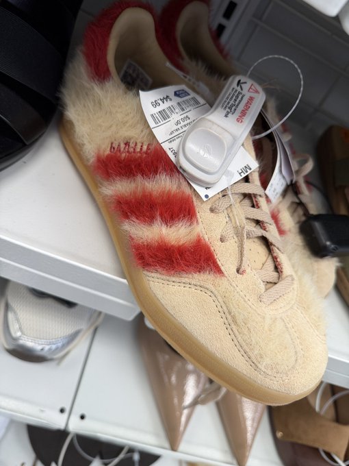 A close-up of Adidas Samba sneakers in cream suede with red furry stripes, complete with gum soles and retail tags still attached, displayed on a shoe shelf.