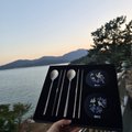 A scenic view at dusk or dawn showing a person's hand holding a small open case containing silver utensils against a backdrop of water, distant mountains, and a sunset sky.