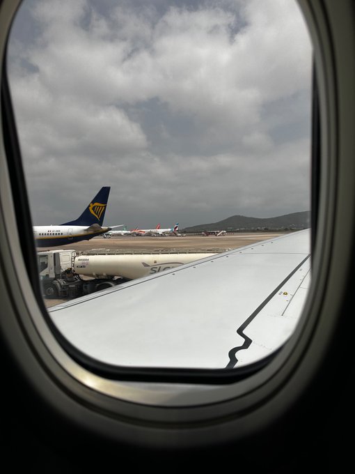 A view through an airplane window shows the wing of the aircraft and several Ryanair planes parked at the gate, including a fuel tanker labeled "FUEL SERVICES" beneath a cloudy sky—capturing the moment of departure for a weekend trip home.