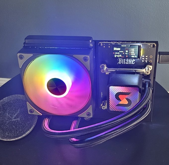 A Bitaxe Bitcoin miner displays a real-time hashrate of 84.04 on its small screen while its RGB cooling fan glows with rainbow colors. The compact device, branded with "Bitaxe," sits alongside a hockey puck for scale, illustrating the small but capable nature of modern home mining hardware.