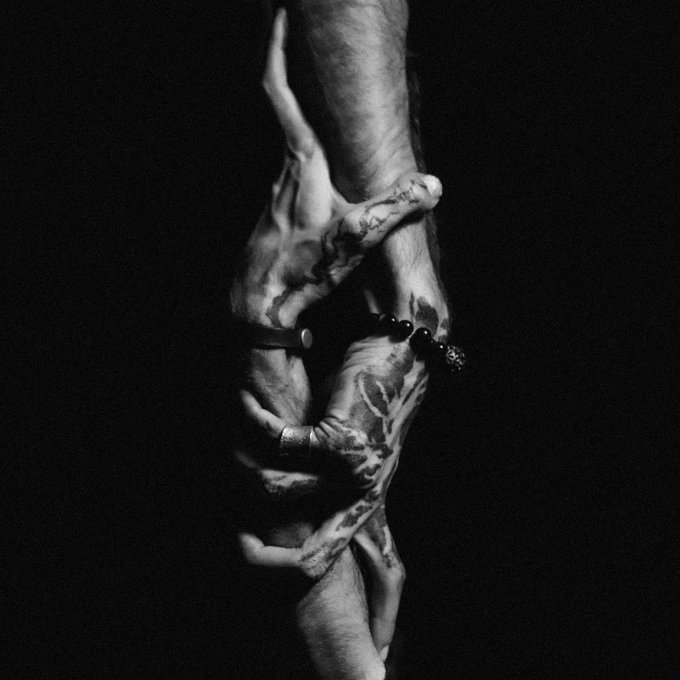 A high-contrast black and white photograph captures intertwined tattooed hands gripping a wrist, with one person wearing a dark beaded bracelet.
