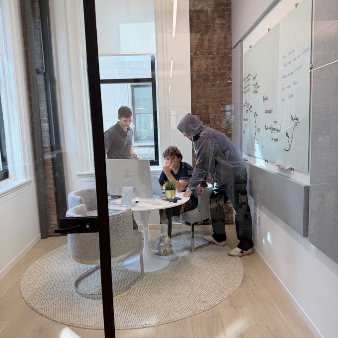 Three young men collaborate around a small table with a laptop and papers in a modern glass-walled meeting room, where the adjacent whiteboard displays extensive brainstorming notes and diagrams likely outlining the "SideShift V3" product roadmap teased in the post.