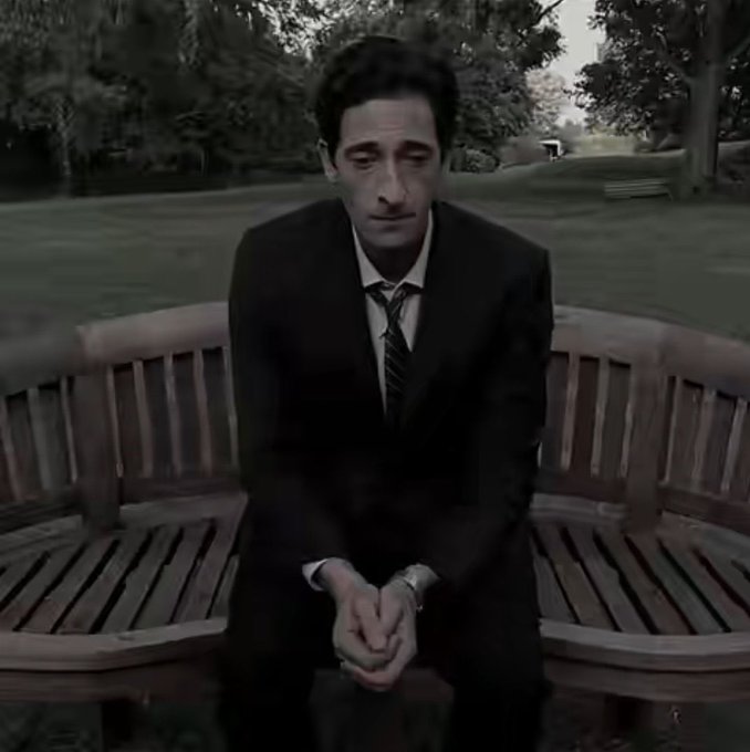 A desaturated, almost black-and-white shot of a man in a dark suit and tie sitting alone on a curved wooden park bench, looking down with a somber expression.