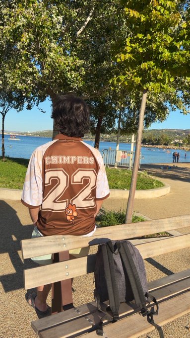 A person wearing a brown and white "CHIMPERS 22" jersey, sitting on a wooden bench with a backpack beside them, looking out at a body of water.