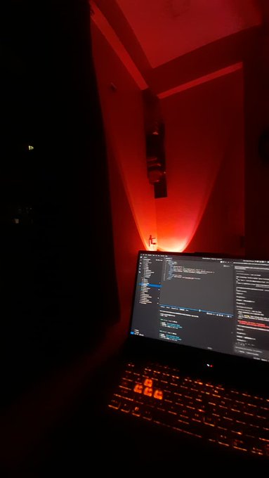 A dimly lit scene featuring an open laptop with code displayed on the screen and an orange-backlit keyboard, illuminated by a glowing red light source in the background.