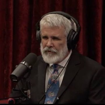 A bearded man with gray hair wearing a suit and floral tie speaks into a microphone while wearing headphones, seated in front of a red curtain.