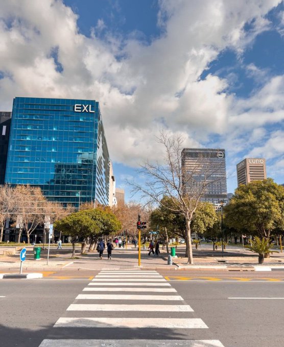 A city street view featuring a crosswalk leading toward modern office buildings, including one prominently displaying the 'EXL' logo and another with 'Luna' signage under a partly cloudy sky.