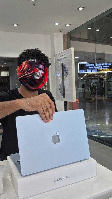 A person wearing a red metallic mask stands in a brightly lit store, holding an open silver MacBook Air above its white product box, with a display poster visible in the background.