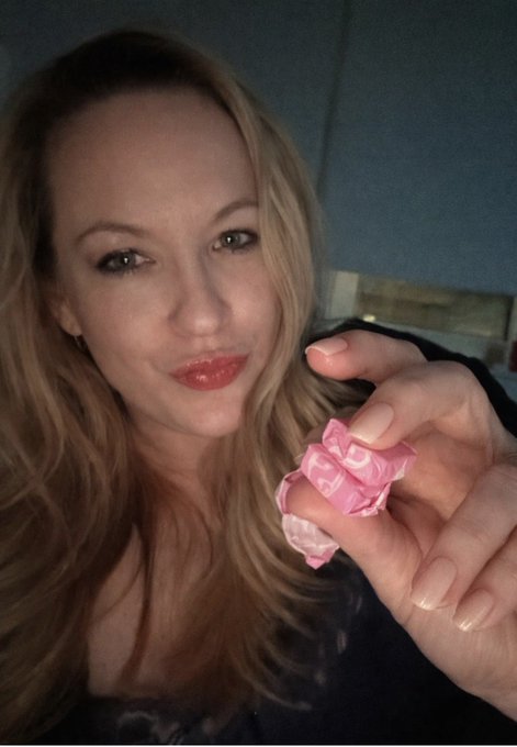 A selfie of a blonde woman with red lipstick holding a pink rose toward the camera.