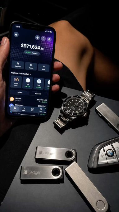 A person's hand holds a smartphone displaying a crypto portfolio totaling '$971,624.18,' resting on a surface alongside several Ledger hardware wallets, metal security keys, and a luxury wristwatch.