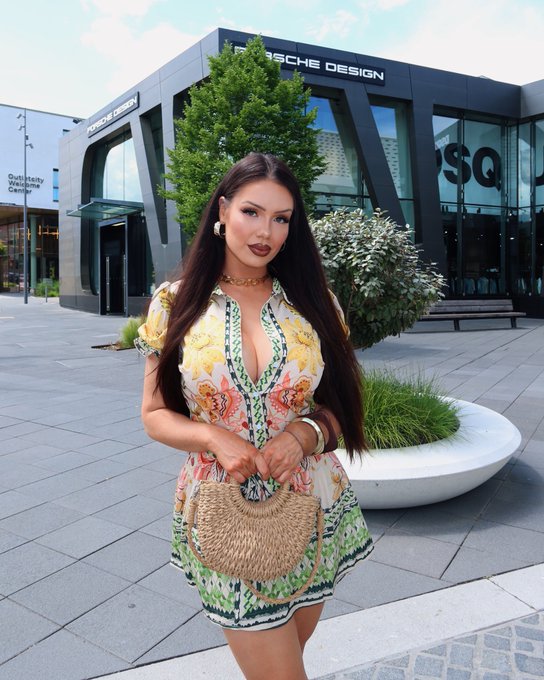 A woman wearing a floral dress stands in front of a Porsche Design store, with visible signage including "Porsche Design" and "GUTHARY Wellness Center." She holds a woven handbag, complementing the upbeat, optimistic tone of the "Grand rising" greeting for a bullish week ahead, with the luxury brand backdrop reinforcing a sense of prosperity and new beginnings.