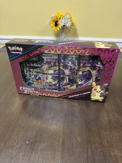 A Pokémon Crown Zenith Morpeko V-Union premium figure collection box sits on a dark wooden table, featuring vibrant pink and purple packaging that displays the Morpeko character and its V-Union cards. The caption "Bad decision time" humorously foreshadows an impulse purchase of this collectible, as the unopened box beckons from the table beside a vase of sunflowers.