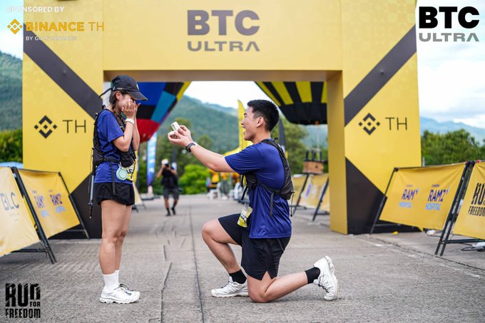 A man kneels with a ring at a "BTC Ultra" finish line as a surprised woman covers her mouth, while sponsor banners for "Binance TH," "Kam," and "Run for Freedom" frame the scene—capturing the Bitcoin event proposal announced in the post.