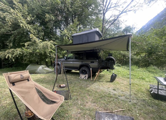 An outdoor overlanding campsite featuring a Toyota Land Cruiser with a roof top tent and awning, a camping chair, a small tent, and a campfire setup in a wooded area.