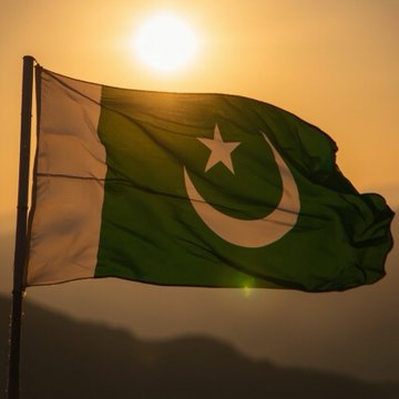 The flag of Pakistan (green with a white crescent and star) waving in the wind against a bright sun.