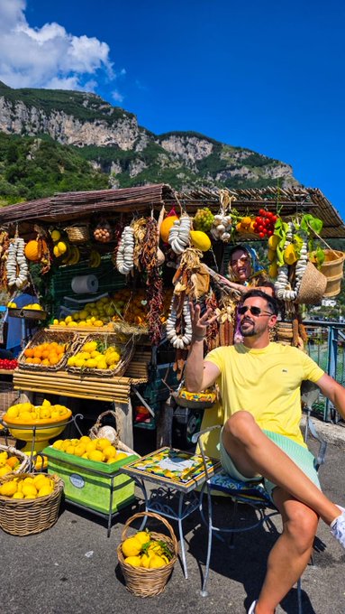 A man in a yellow shirt and green shorts sits smiling at an outdoor fruit market stall filled with lemons, peppers, and other produce, with mountains in the background.