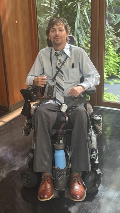 A man in a wheelchair wearing a dress shirt, tie, and lanyard, with a water bottle attached to his chair, positioned indoors near plants.
