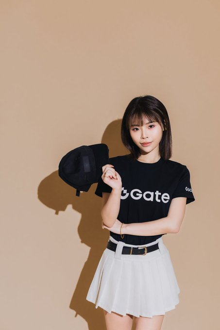 A young woman with short dark hair poses against a tan background, wearing a black t-shirt with white "Gate" text and a white pleated skirt while holding a black cap.