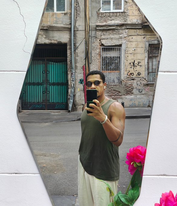 A mirror selfie of a muscular man wearing sunglasses, a dark tank top, and white pants, standing on a street with old buildings in the background.