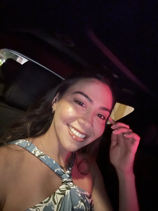 Smiling young woman taking a selfie in a dimly lit car with pink/red ambient lighting, wearing a blue and white patterned halter top.