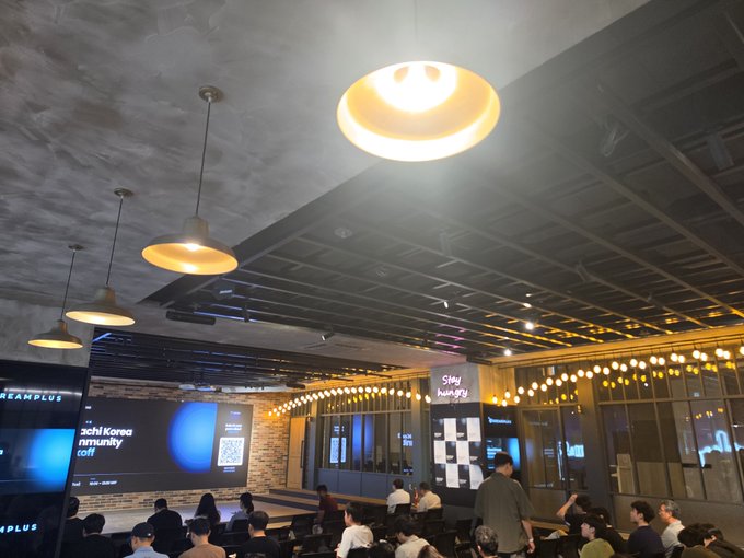 A well-lit event space with industrial-style ceiling and hanging pendant lights hosts the "Hibachi Korea Community" kickoff meetup, where attendees are gathered in front of a stage displaying the event branding alongside a DreamPlus logo, capturing the networking atmosphere described in the post.
