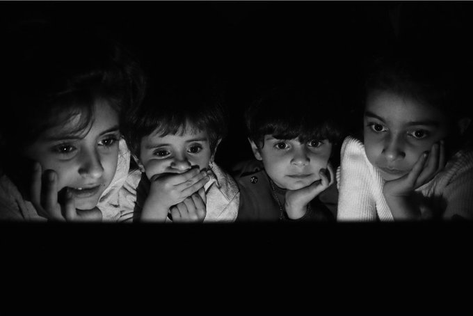Black and white photograph of four children with somber expressions peering out from the darkness, each resting their chin on their hands.
