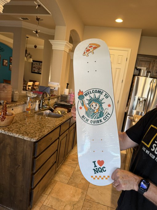 A person holds a white skateboard deck featuring a pizza slice graphic, a 'Welcome to New Quirk City' Statue of Liberty emblem, and the text 'I ❤️ NQC' with a checkered border.