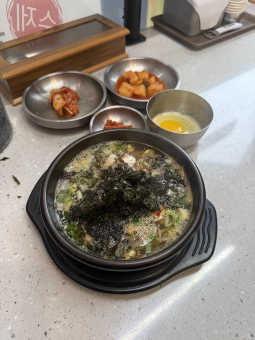 A bowl of Korean soup topped with seaweed and green onions is surrounded by side dishes including kimchi, radish, and a raw egg.