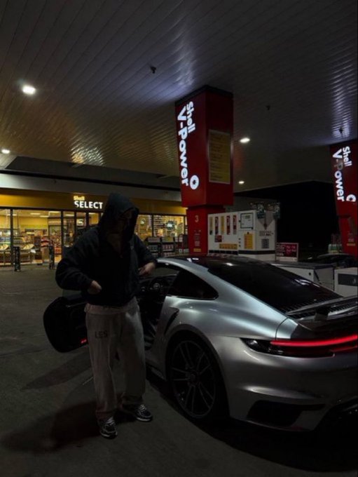 A nighttime photo of a person in a black hoodie standing next to a silver Porsche 911 at a Shell gas station.