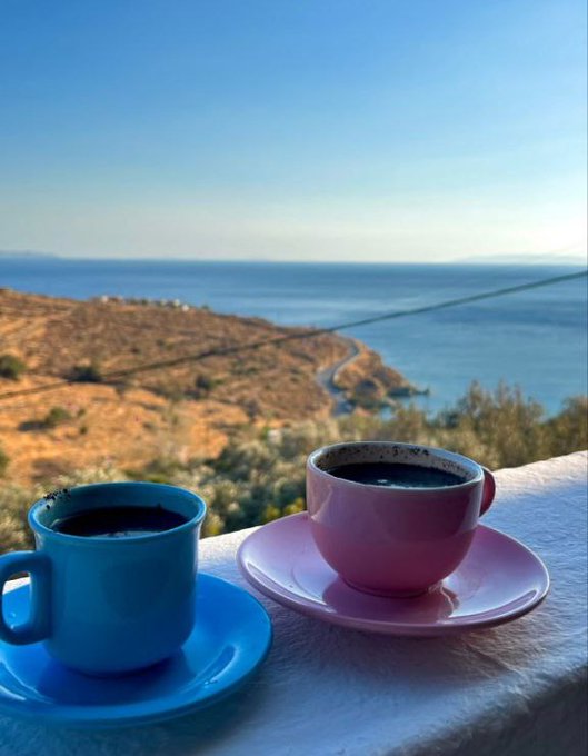 A scenic photograph of two coffee cups (blue and pink) on saucers placed on a white table, overlooking a beautiful ocean and coastal landscape.