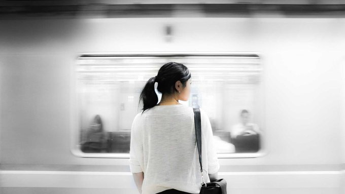 A woman with a ponytail stands on a train platform, gazing at a passing train whose motion blur contrasts with her still figure. The image captures the tension between waiting and movement, mirroring the post's reflection on how immature markets overreact to policy shifts rather than steadying themselves with patience.