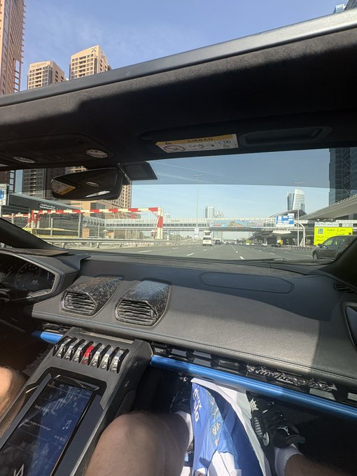 The interior of a luxury sports car (likely a Lamborghini Huracán, given the distinctive center console toggle switches) is shown driving on a city highway under bright blue skies, with the passenger displaying a "GM" graphic on their shirt. The image relates to the post's "GM" text through the visible initials, which may reference "General Motors" or stand for something else entirely in this context of exotic automotive enthusiasm.