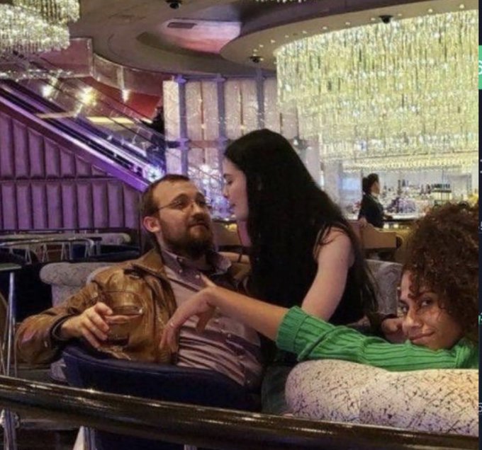 The image features an indoor setting with three people sitting around in chairs. One person is holding a glass of wine while another person is petting them, possibly a woman. A third person can be seen nearby watching this interaction. In addition to these individuals, there are also two more chairs visible in the scene. The presence of multiple chairs suggests that it could be a restaurant or an upscale venue where people gather for socializing and enjoying drinks.
