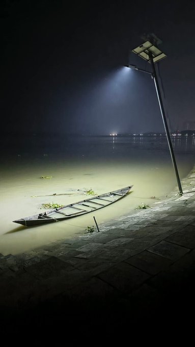 A nighttime photograph of a wooden canoe resting on the muddy shore of a body of water, illuminated by a single bright streetlamp that casts a dramatic beam through the dark, misty atmosphere.