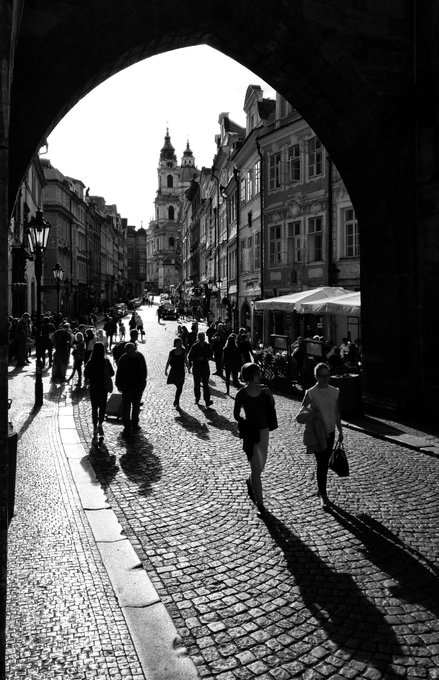 A black-and-white photograph taken through a stone archway shows people walking along a cobblestone street, with a Gothic church tower and a church spire visible in the distance.
