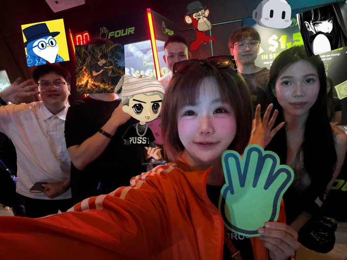 A group of young people take a selfie at a promotional event, with one woman in an orange jacket in the foreground holding up a green foam hand, while cartoon character cutouts decorate the background.