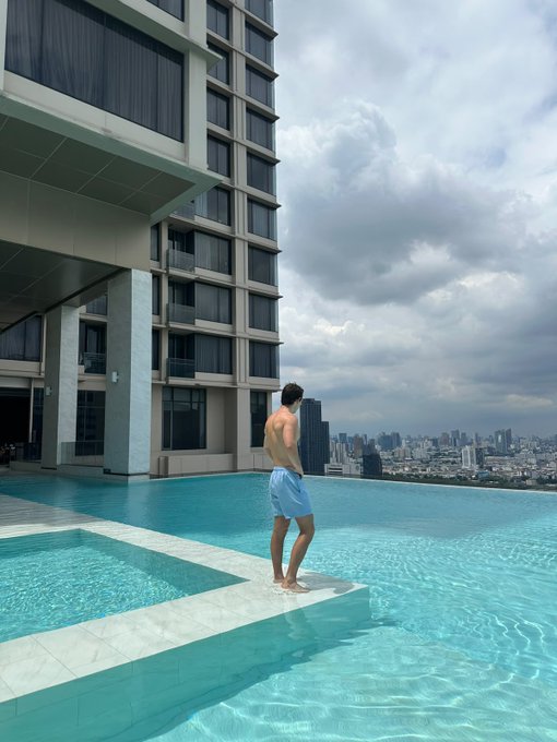 A shirtless man in light blue shorts stands at the edge of an infinity pool on a high-rise building, overlooking a sprawling cityscape under a cloudy sky.
