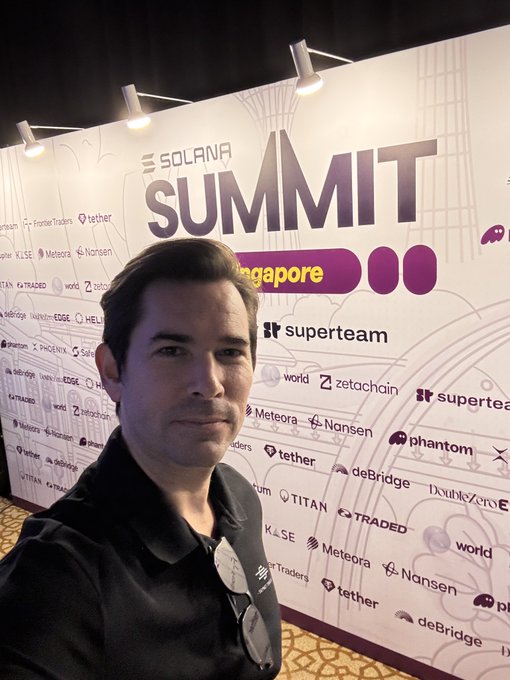 A man wearing a black polo shirt takes a selfie in front of a 'Solana Summit Singapore' backdrop featuring various sponsor logos including Tether, Phantom, and Superteam.