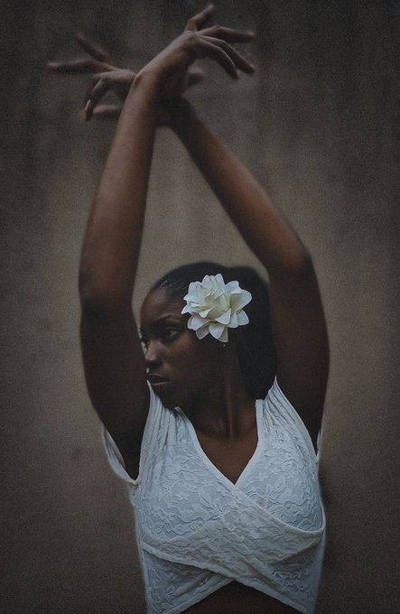 A moody portrait of a dark-skinned woman in a white lace top with a white flower in her hair, arms raised gracefully overhead against a textured brown background.