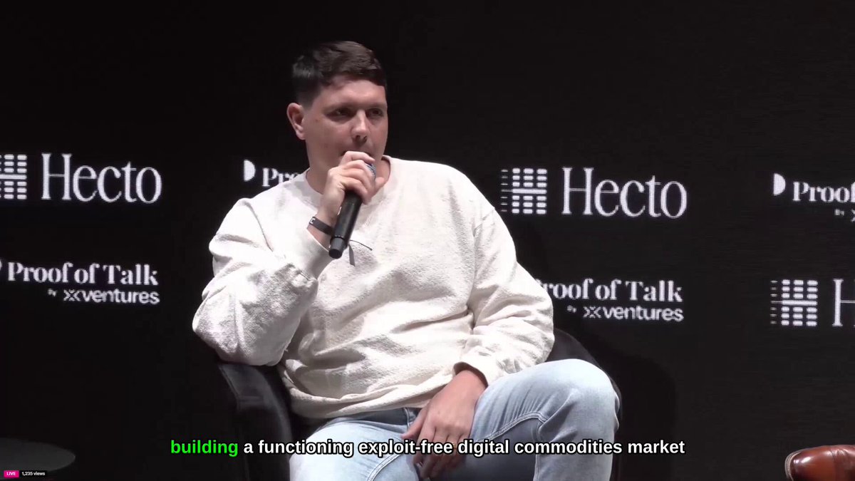 The image shows a man sitting down with his hands on his lap while holding a microphone, possibly speaking at an event or giving a presentation. The background features a black wall with white text that reads "Building a functional exploration free digital commodities market". This suggests that the man might be discussing the concept of Bittensor subnets and their importance in the context of the digital commodities market.
