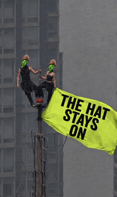 Two performers in green vests balance atop a city lamppost, unfurling a bright yellow banner reading "THE HAT STAYS ON," visually embodying the bold and attention-grabbing branding of the $RobinWifHat crypto project being promoted.