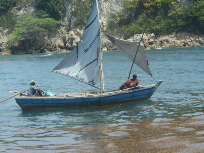 Two people in a small blue sailboat with a tattered white sail on turquoise water, with a rocky, green-covered coastline in the background.