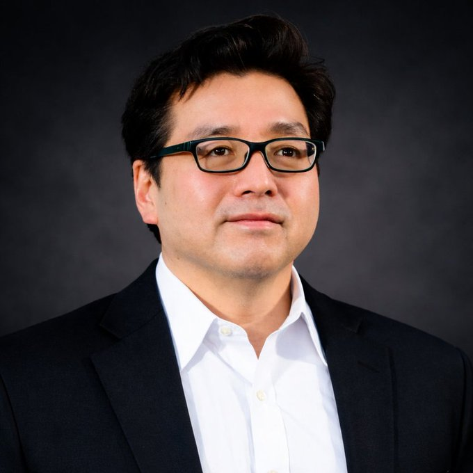 A headshot portrait of an East Asian man with glasses, wearing a black suit jacket and white dress shirt against a dark grey background.