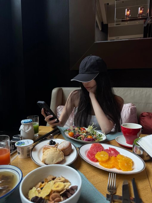 A woman in a black cap is seated at a breakfast table, surrounded by plates of fresh fruit, pastries, salad, and granola while looking down at her phone.

The image shows a woman casually scrolling on her device amid a leisurely meal—an everyday moment of disengagement that contrasts with the post's discussion of @vangrid_io, a platform built around active, intentional engagement where users fund spatial data requests and contributors compete to fulfill them.